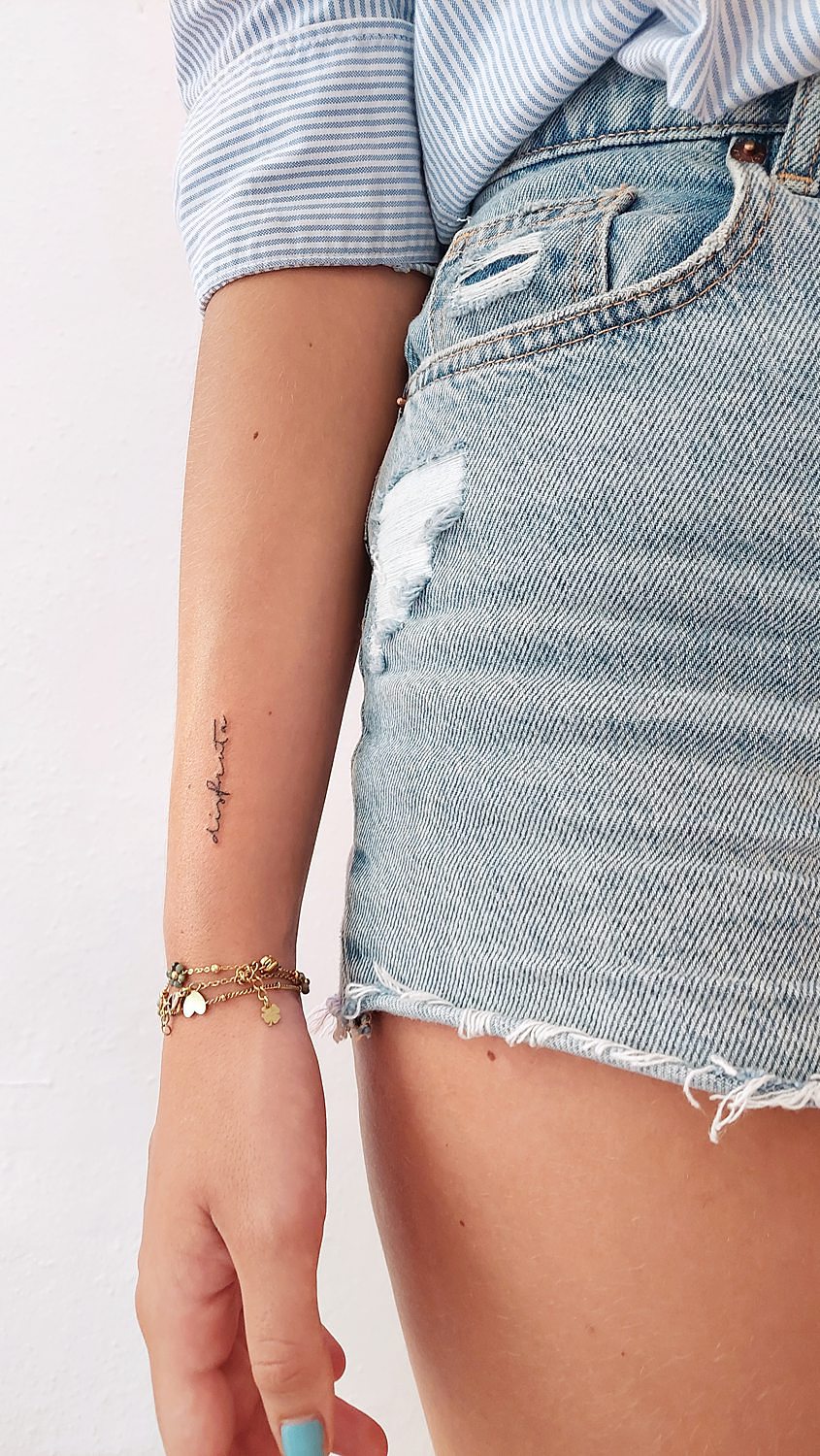 Tatuaje brazo con la frase "disfruta" escrita en cursiva y línea fina