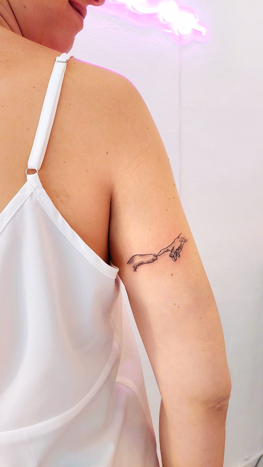 Tatuaje minimalista de mano y pata de perro simulando el cuadro de La Creación de Adán en brazo derecho
