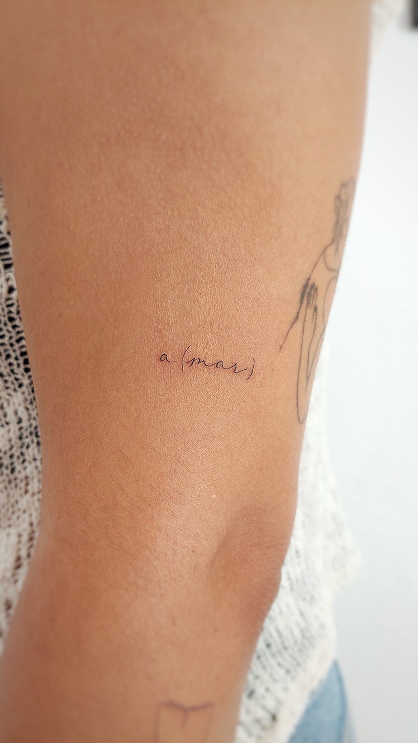 Tatuaje minimalista en el brazo con texto 'a(mar)'.