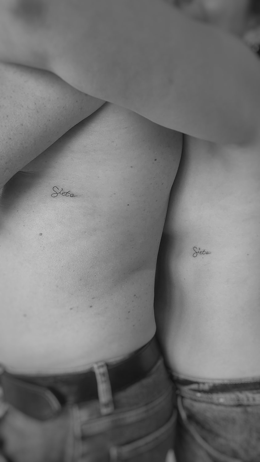 Two people with matching "Siete" tattoos on their torsos.