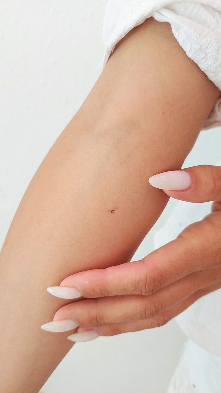 Close-up of a tiny lowercase 'i' tattoo on an arm.