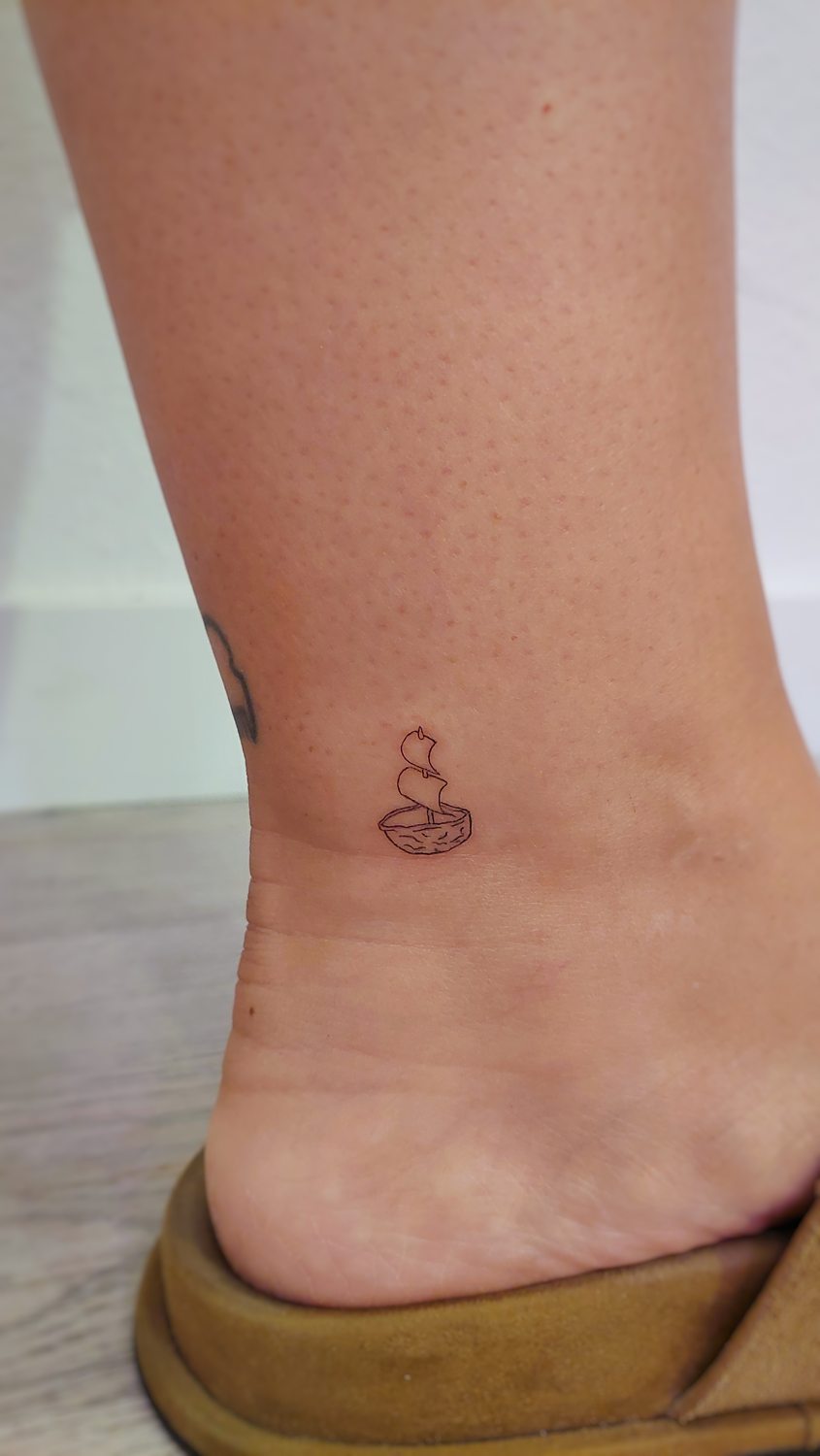 Tatuaje minimalista de un barco de vela en el tobillo.